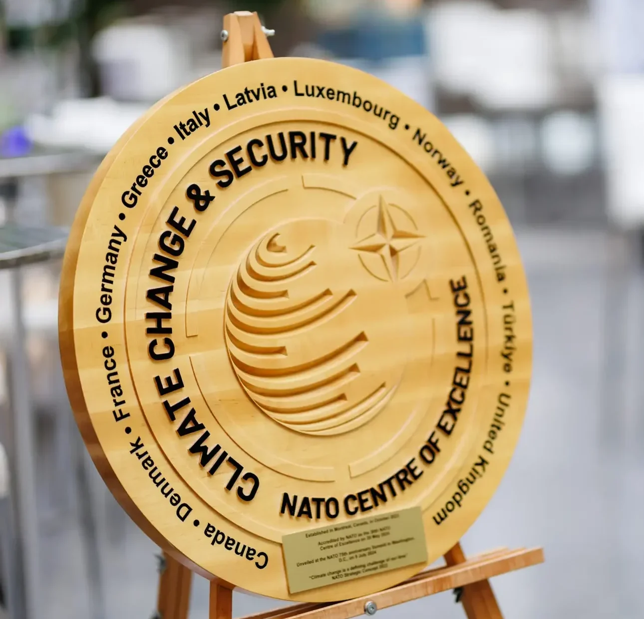 Wooden CCASCOE emblem on an easel featuring the text ‘Climate Change & Security – NATO Centre of Excellence,’